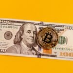 Gold bitcoin placed on a 100 US dollar bill over a bright yellow backdrop, symbolizing modern digital currency.