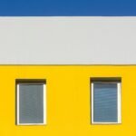 Minimalist architecture featuring a vibrant yellow facade with two windows and a blue sky.